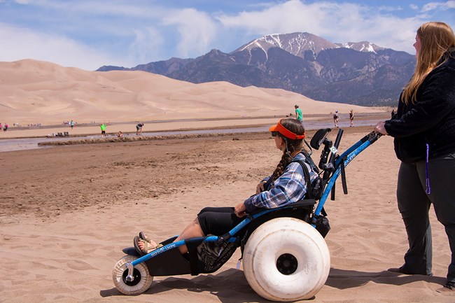 A woman pushing another woman in a newer style dunes wheelchair with Medano Creek, dunes, and a mountain in the background