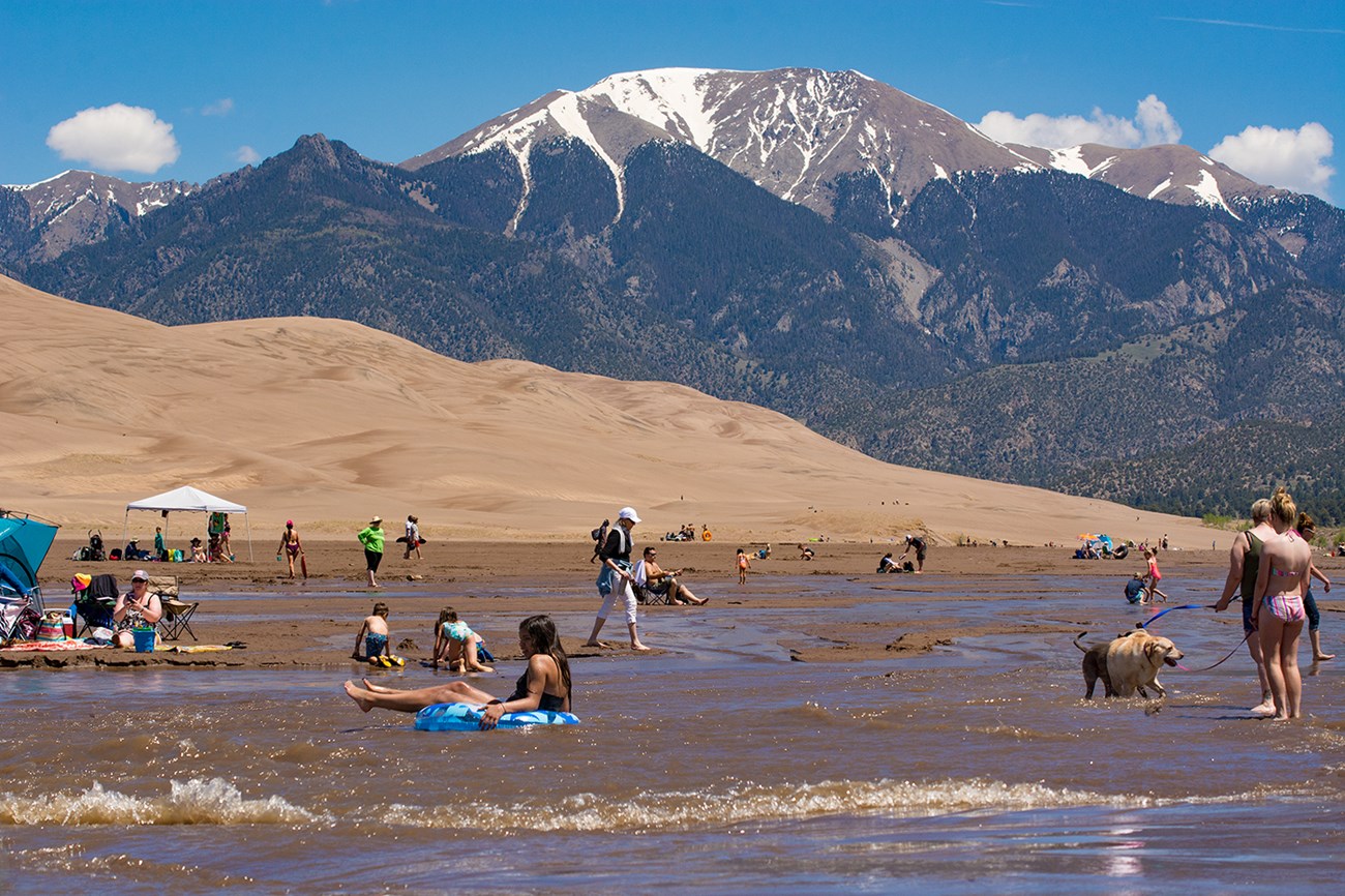 A wide creek with small waves, visitors playing in the water, dunes and snow-capped mountain in the background