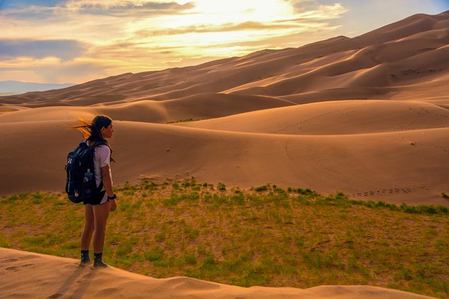 A teen girl pauses to take in the view of dunes illuminated at sunset as she hikes up a dune slope