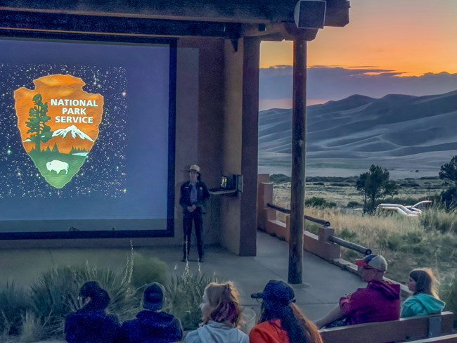 A ranger stands in front of a screen at the amphitheater at sunset, starting an evening program