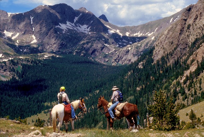 Two people sit on horses at Music Pass, with an alpine basin of trees and tundra with patches of snow