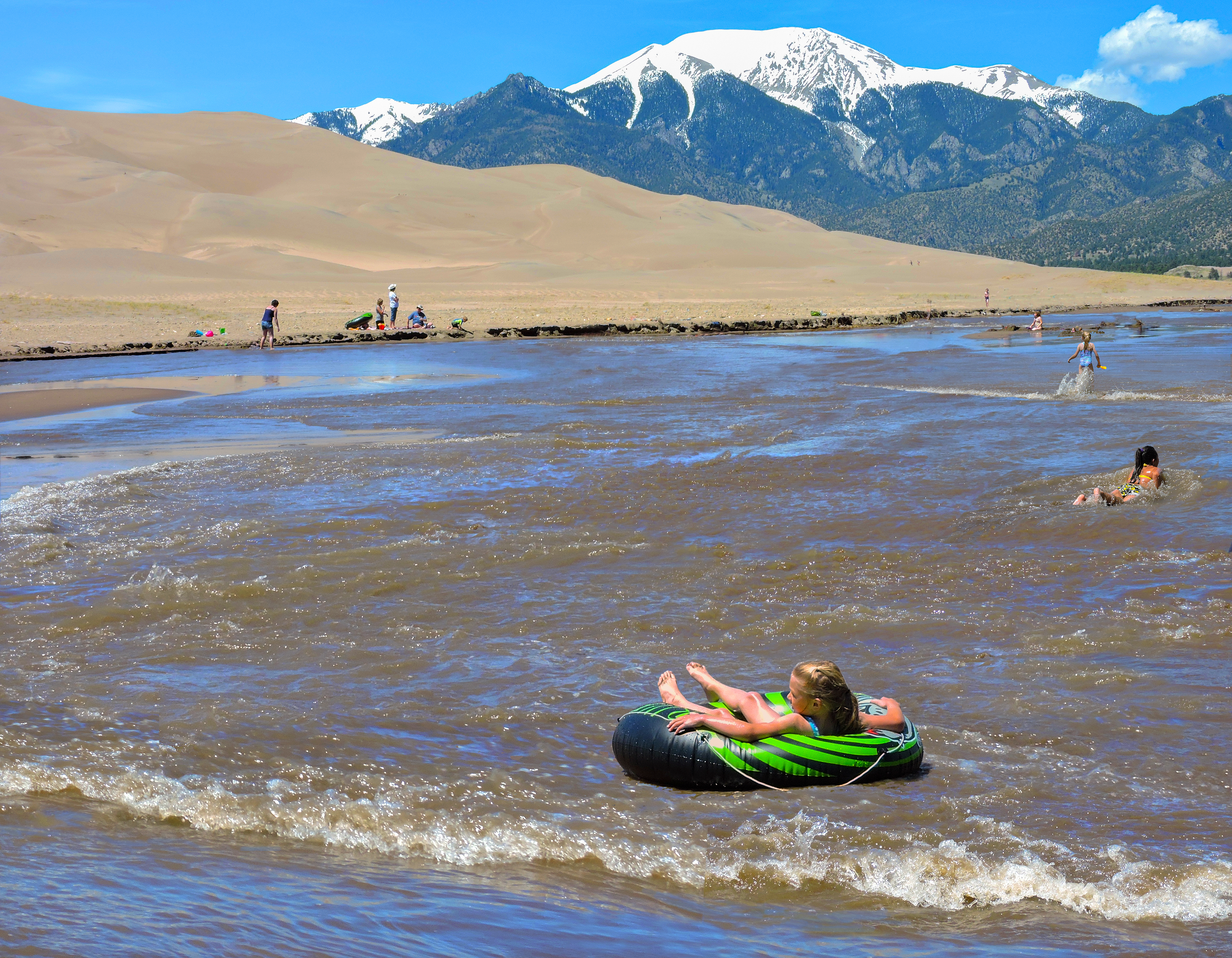 A girl in the foreground floats on a tube in a wave of Medano Creek, while other youth splash and play. In the background are dunes and a snowy mountain.