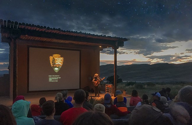 A ranger plays guitar to open an evening program in front of an audience at the outdoor amphitheater. The dunes are silhouetted under a sky with clouds and stars.
