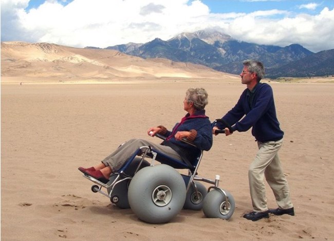 A man pushes a woman in a dunes wheelchair at the base of the dunes