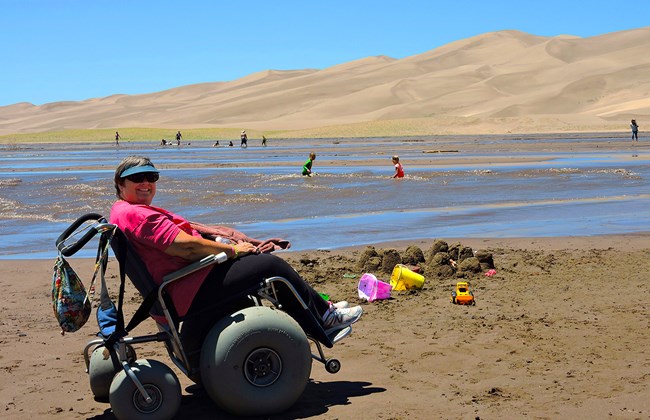 A woman sits in a baloon tire wheelchair on the sand beside a shallow creek and dunes