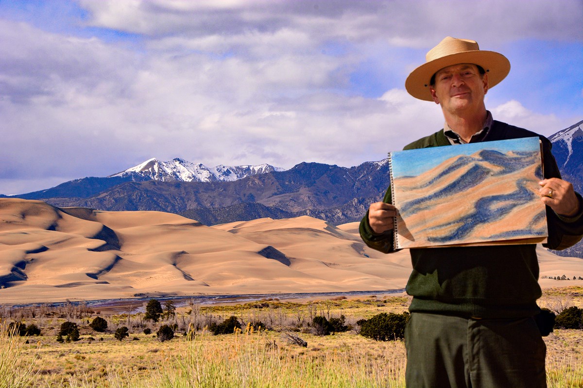 Park ranger holding a color sketch of the dunes, with the dunes and snowcapped mountain in the background