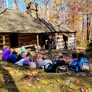students watching a ranger present a program outside a historic cabin in the fall with trees in the background.