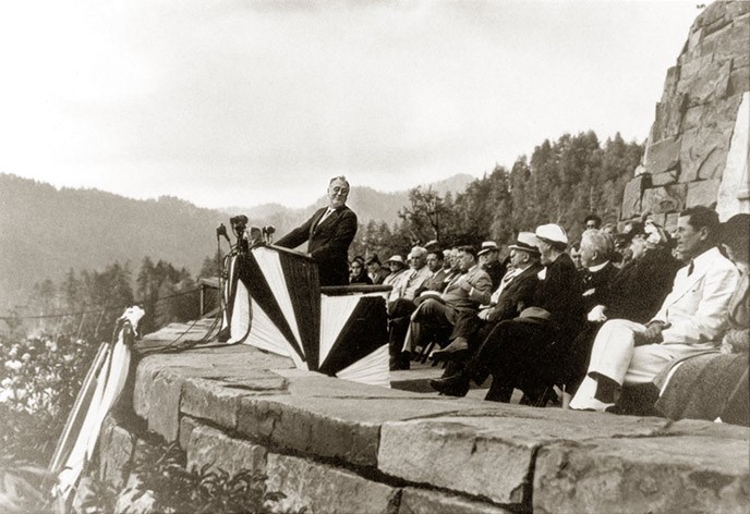 President Franklin Roosevelt stands at the Roosevelt Memorial at the dedication ceremony facing the audience, and a group of people sits behind him.