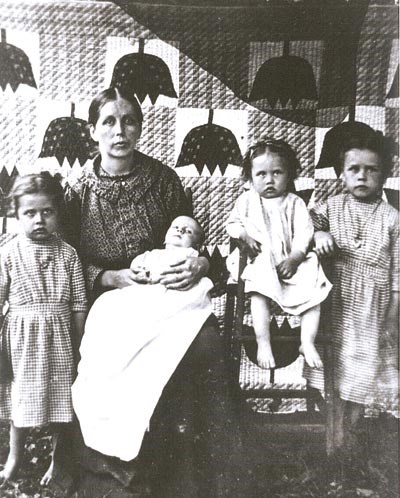 A historic photo of a mountain women posing with her four young children in front of a quilt