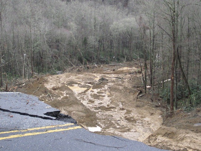 Cracked pavement near rushing, muddy water surrounded bare trees in winter