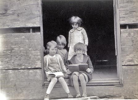 Historic photo of 4 young girls sitting and standing in the doorway of a log schoolhouse
