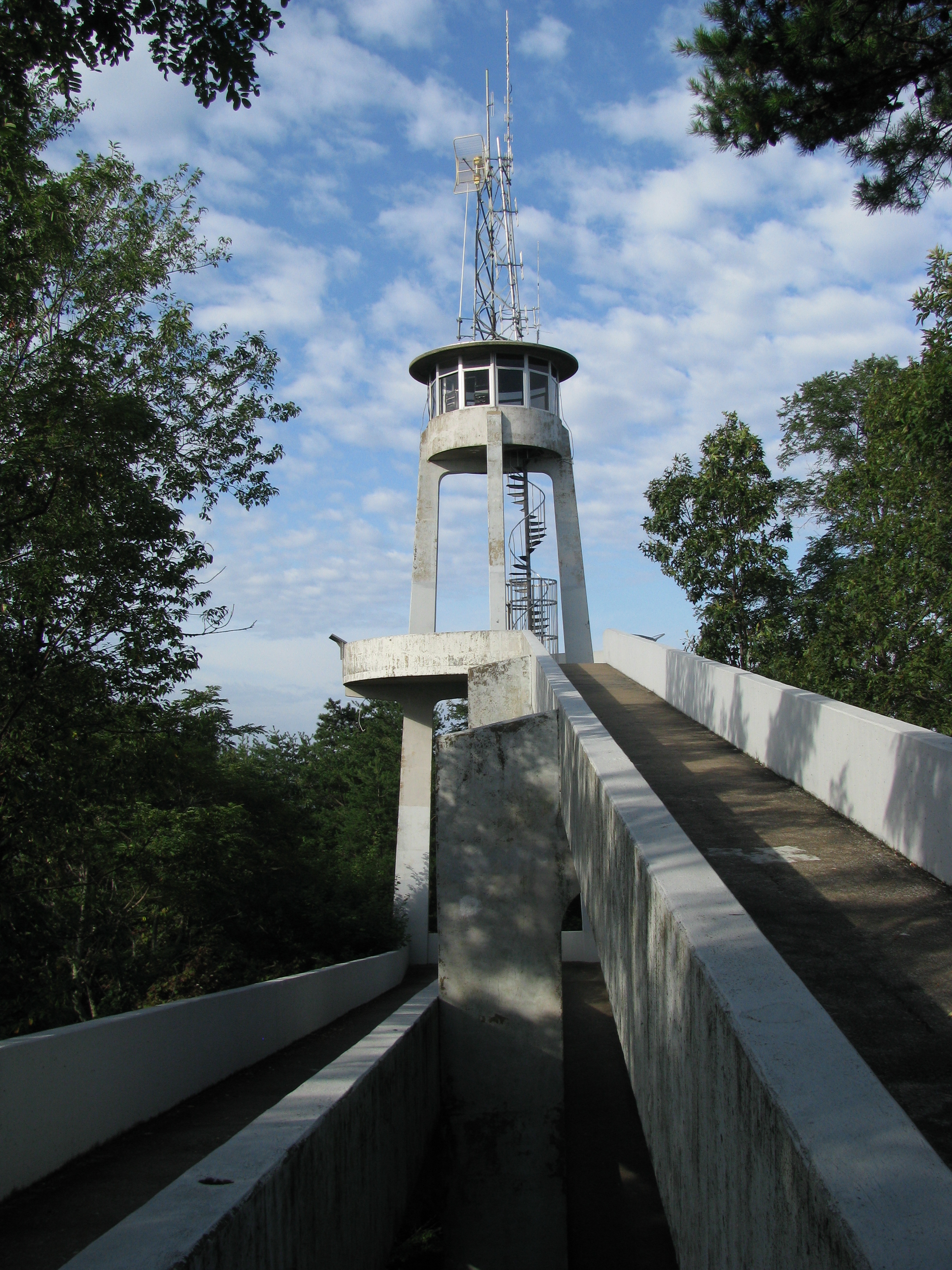 A tower with radio equipment on top of it and a paved path leading up to it. Green trees are seen on either side and a partly cloudy sky is in the background.