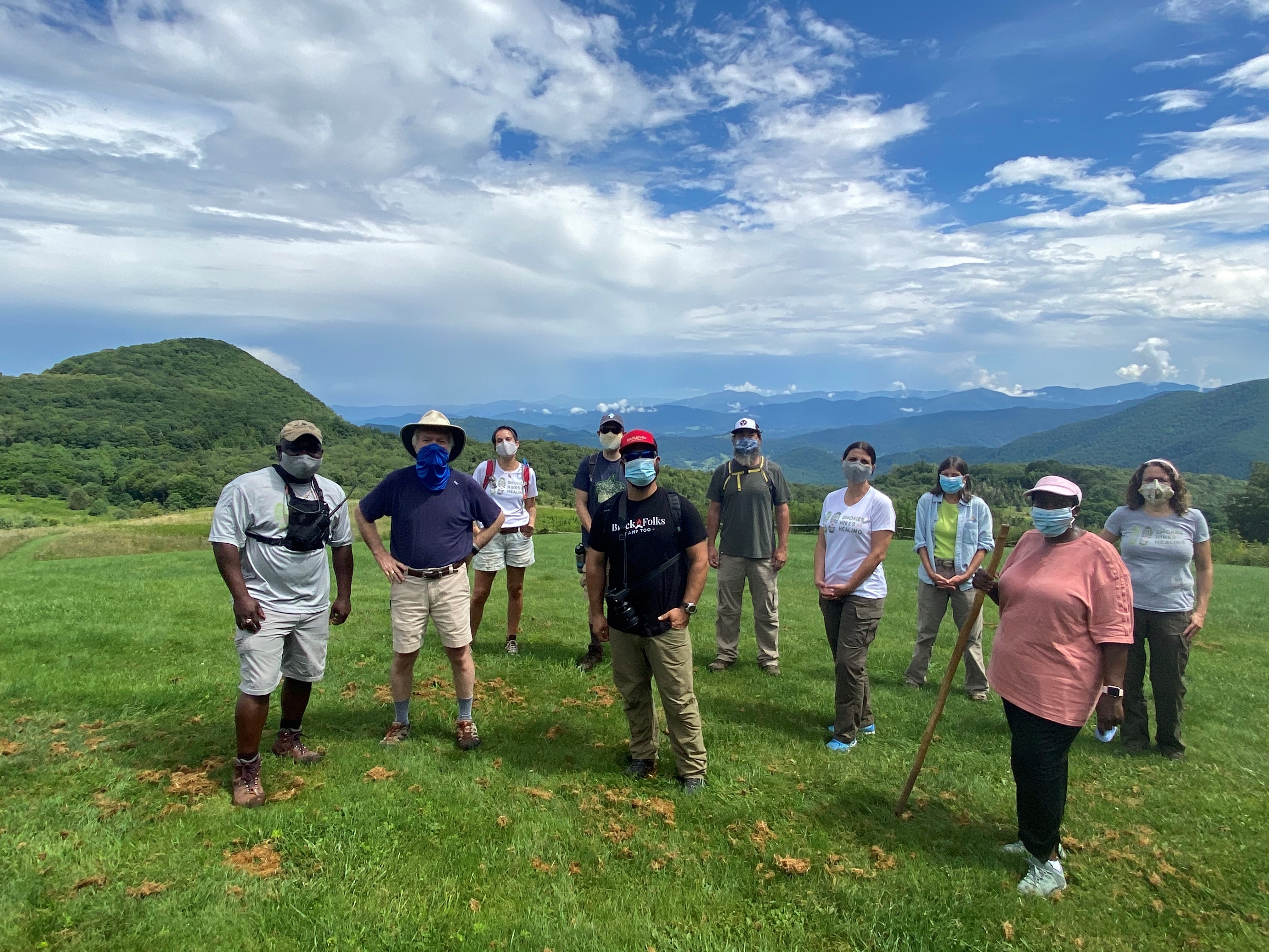 Ten people standing in face masks in the grass with mountains in the background.