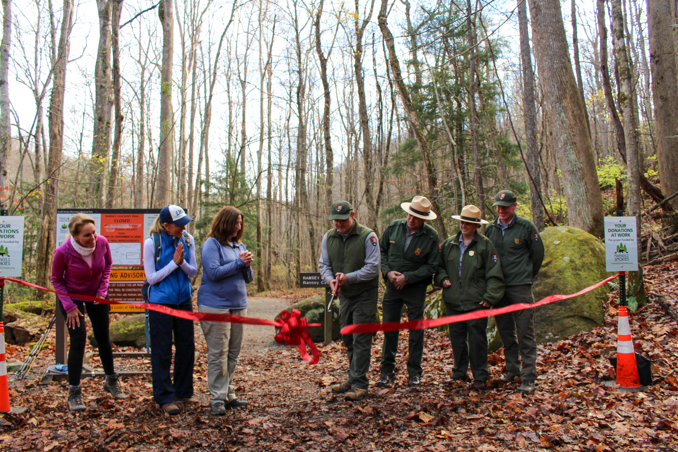 A red ribbon with a bow being cut and falling in front a group of park rangers and Friends of the Smokies staff. Fallen leaves, trees, and a moss-covered rock are in the background.