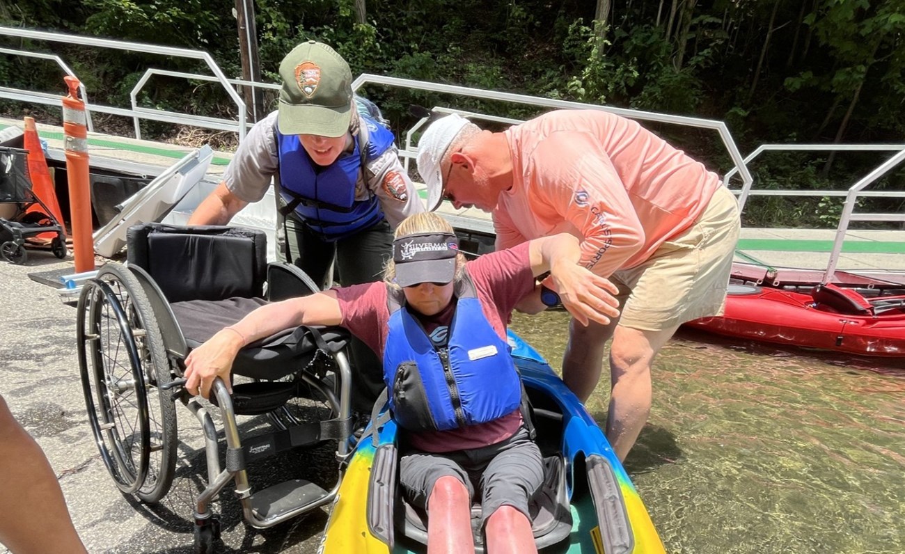 A ranger and another person help someone into a kayak from their wheelchair.