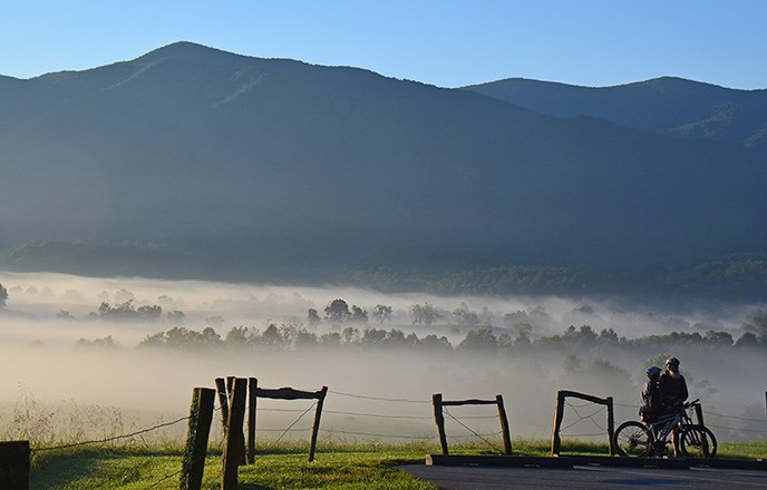 Bicyclists in Cades Cove pause along the road. Behind them mountains rise up from a foggy valley.
