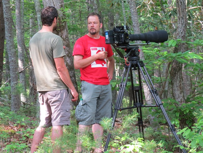 two men standing in the woods behind a camera on a tripod.