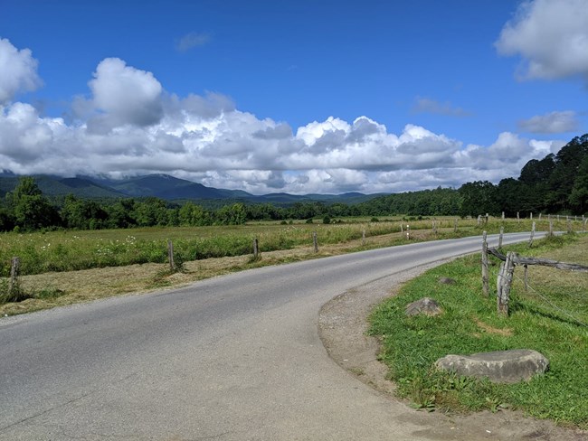 A paved road with a wooden fence and grassy fields on each side. Rolling mountains are in the distance beneath fluffy clouds.
