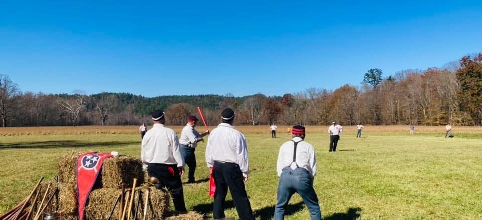 A group of people in a field playing baseball. A stack of hay bales with a Tennessee flag sits near the home plate.