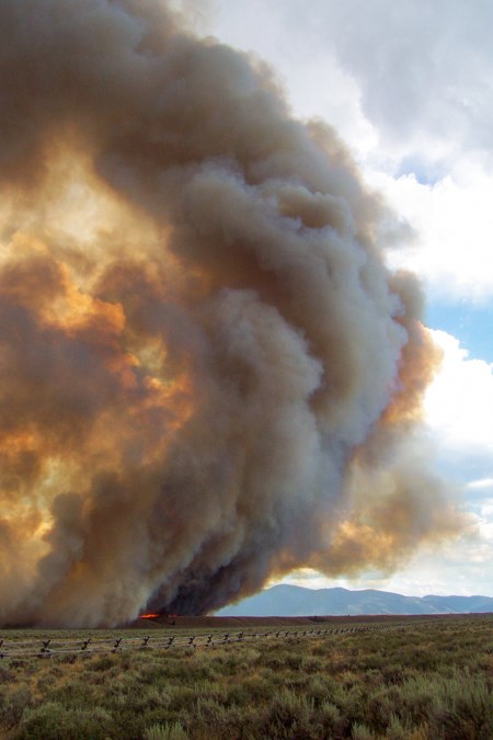 A fire moves across the flat, sagebrush-covered plains, putting up a column of smoke into the air.