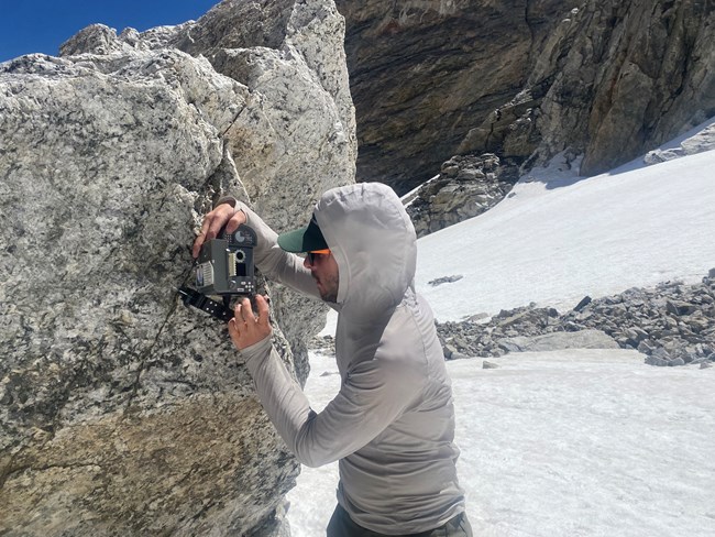 A member of the glacier monitoring crew uses a piton stake to install a timelapse camera onto a boulder