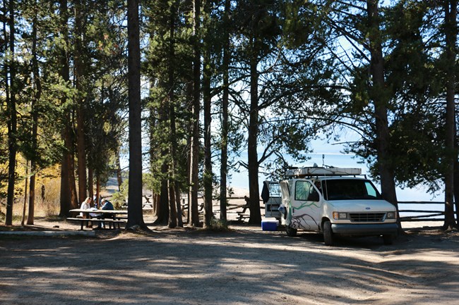 A van parked under trees by a picnic table.