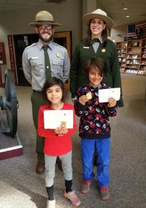 Two park rangers stand behind two new junior rangers holding their badges