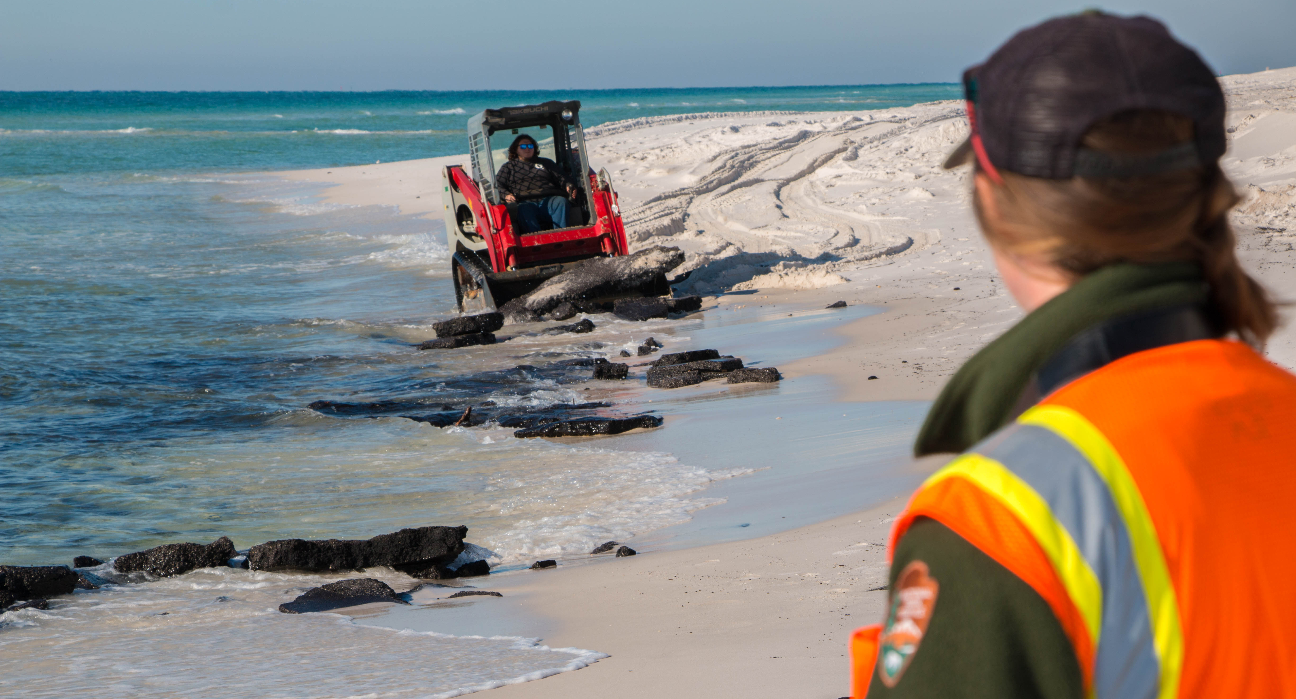 A small front-end loader picks up asphalt debris from the beach at the water line as a ranger looks on.