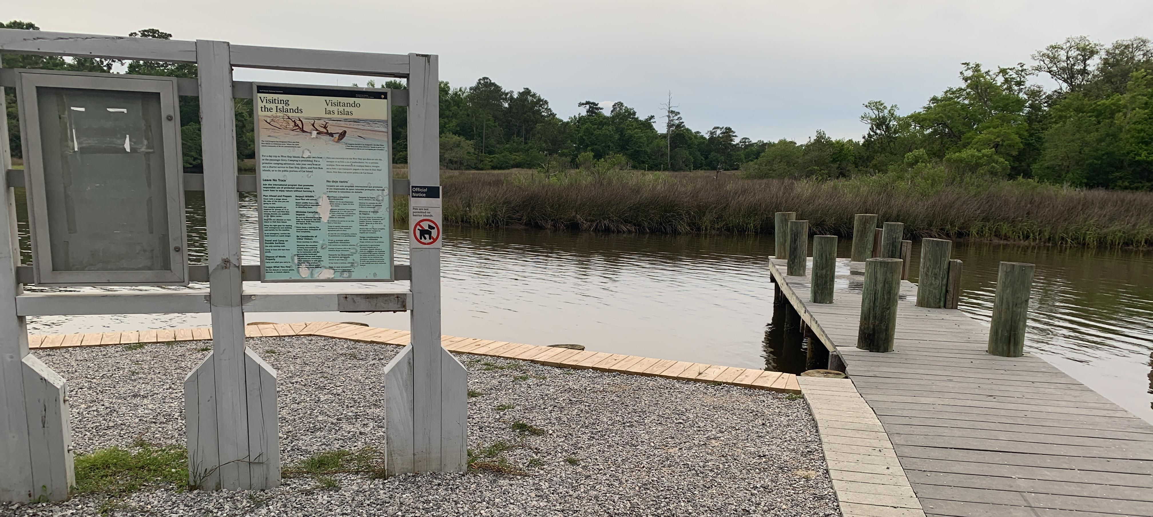 A wooden sign stands in front of a finger pier extending over water in front of salt marsh.