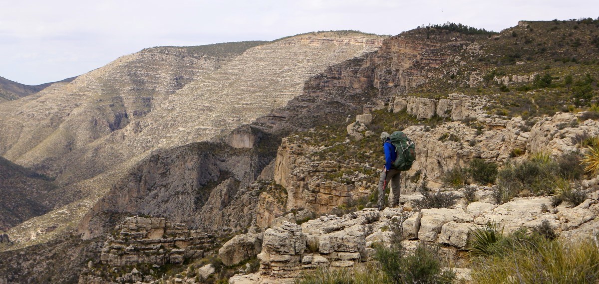 A lone hiker stands at the edge of a great cliff with desert mountains behind him.