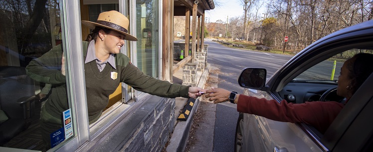 On the left, a park ranger reaches out the window of the fee booth to take a card being handed to her by a visitor, on the right, who is reaching out of the window of her car.