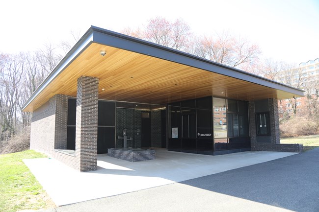 A single-story building with a large awning over benches in front of a drinking fountain and doors to restrooms.