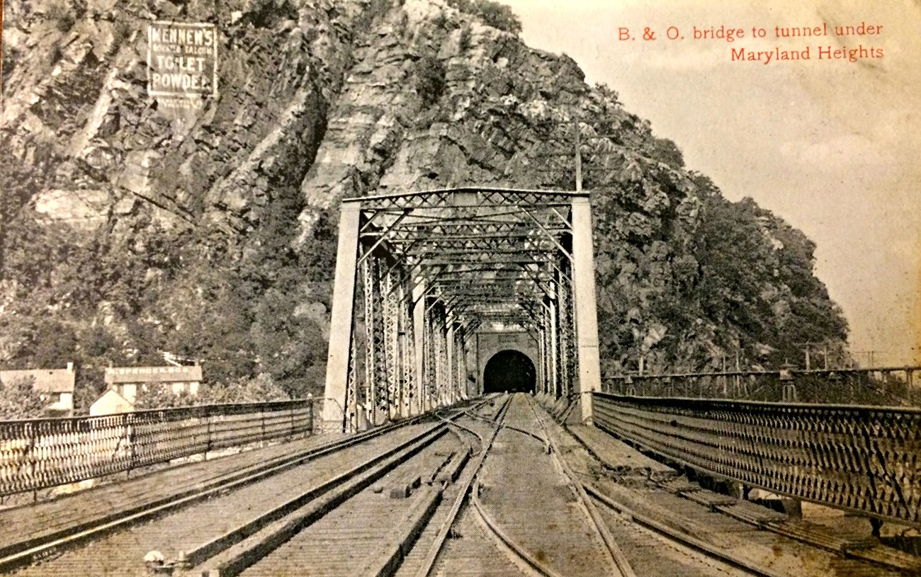 Black and white postcard of white lettered advertisement on cliff face above train tunnel: Mennen's borated talcum toilet powder