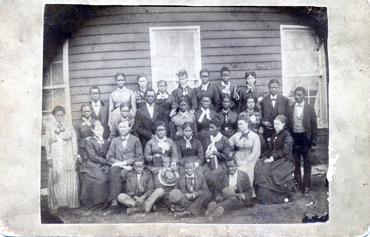 a black and white image of mostly young African Americans seated and standing in front of a board building; white teachers are seated in the group