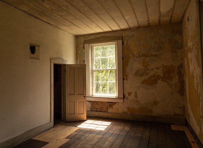 An empty room in a historic building. In the left corner is an open door next to a large window.