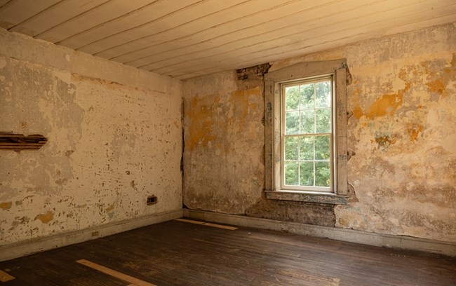 An empty room in a historic building with a large window in the middle right of the photo. There is plaster and paint peeling off exposed brick and wooden floors.