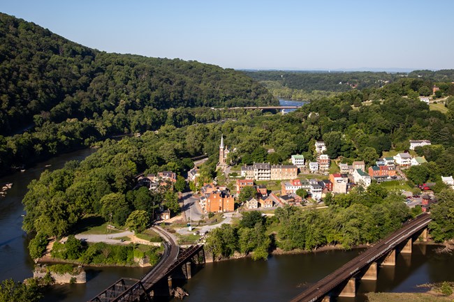 Aerial view of a small town surrounded by trees at the edge of a river with two railroad bridges.