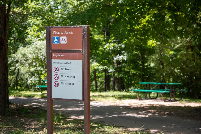 A sign for a picnic area and its regulations next to a picnic table surrounded by trees in the background.