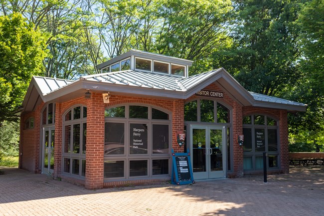 A rectangular brick bulding with windows and doors on three sides surrounded by a paved patio with trees.