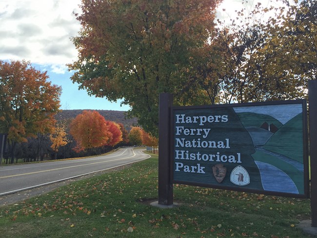 Park entrance sign reads "Harpers Ferry National Historical Park"; it is a painted sign showing a river, church steeple, and mountains. Trees with fall foliage are behind the sign and to the left is a road and more trees