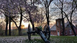 landscape showing flowering trees, an artillery piece, and a brick church; a sunset is in the distance