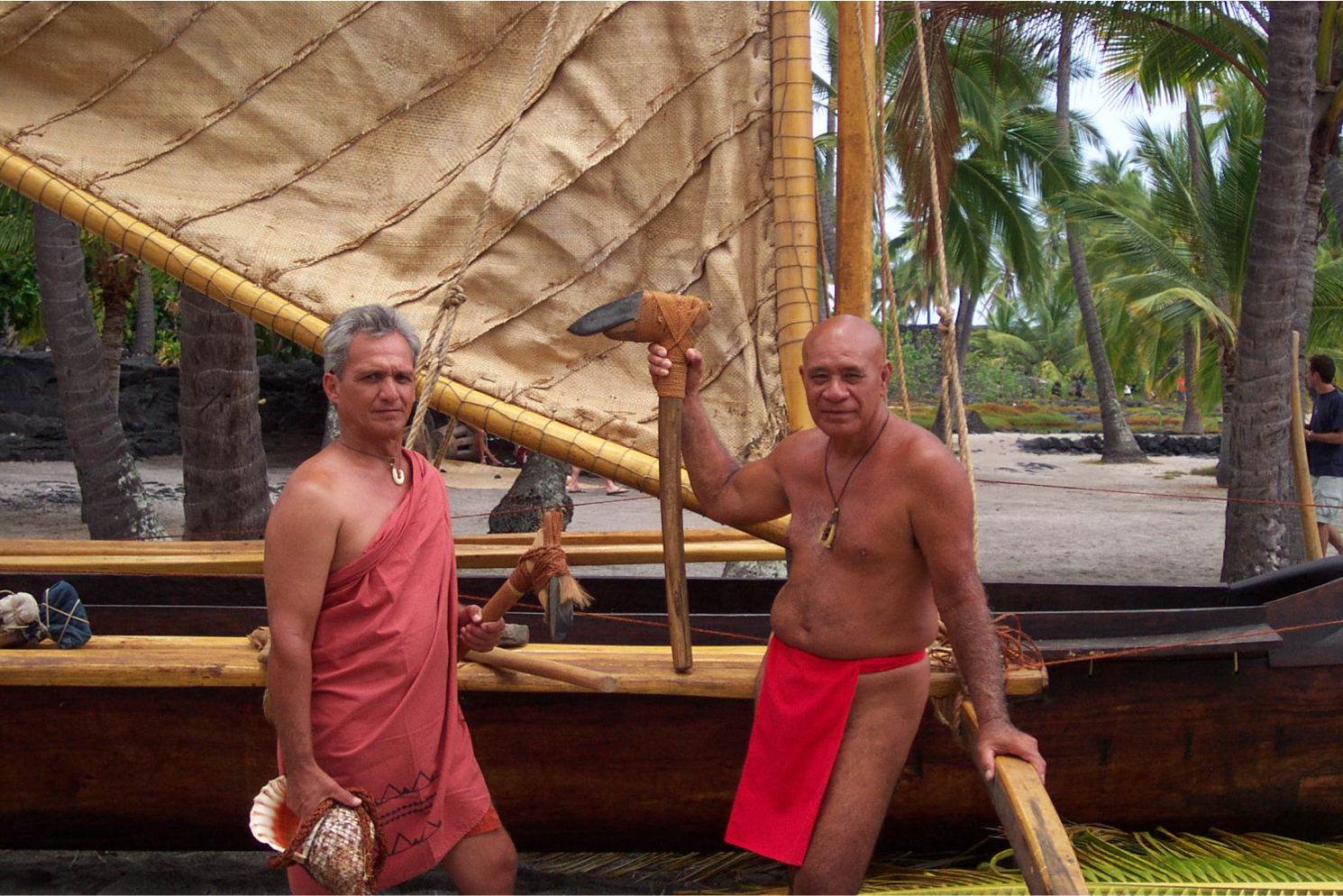two men in traditional Hawaiian dress stand beside hand carved canoe on shore with palms in the background