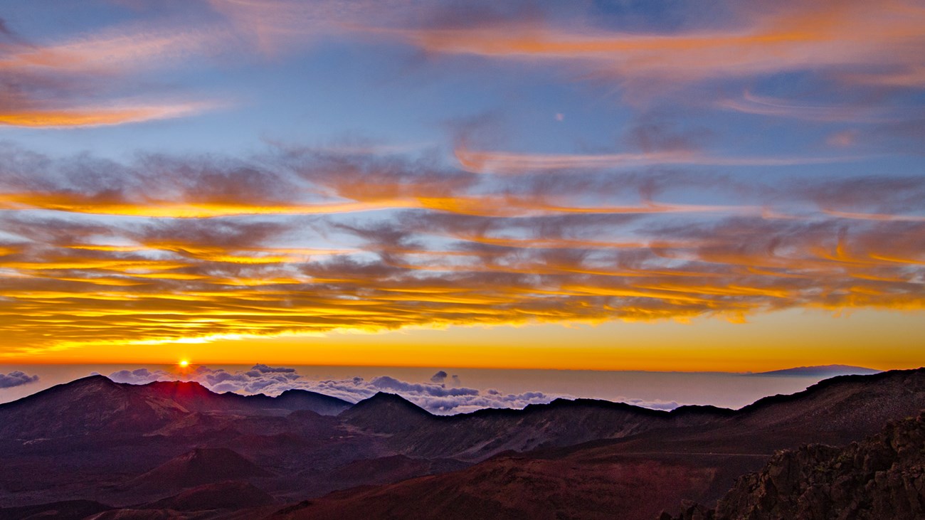 orange and yellow sun rising above the clouds with peaks in the foreground