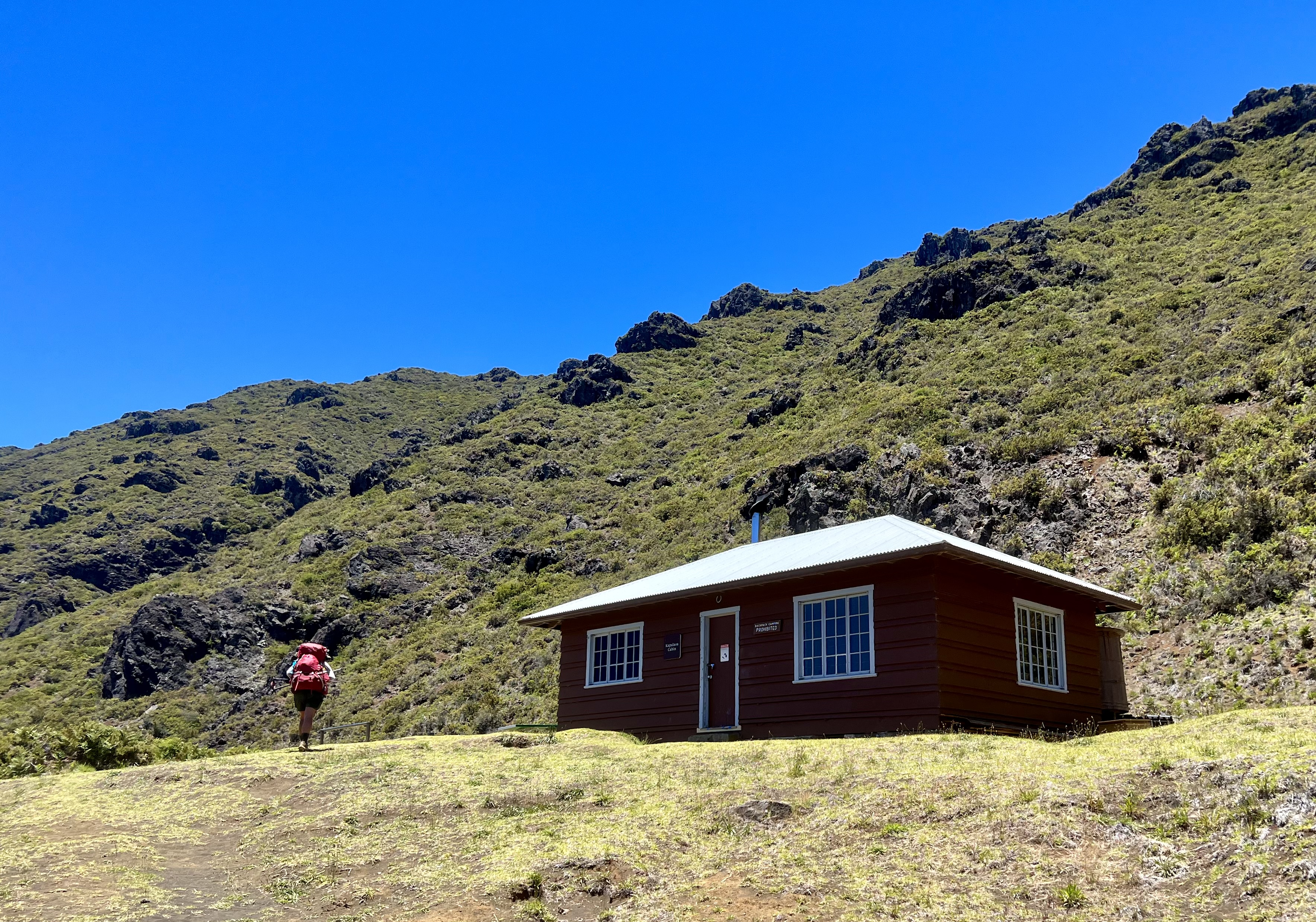 rocky cliff with greenery and cabin in foreground with hiker walking