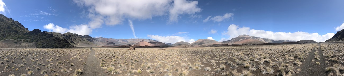 A panorama of a dirt trail extending through a grassland, surrounded by volcanic peaks and cinder cones.