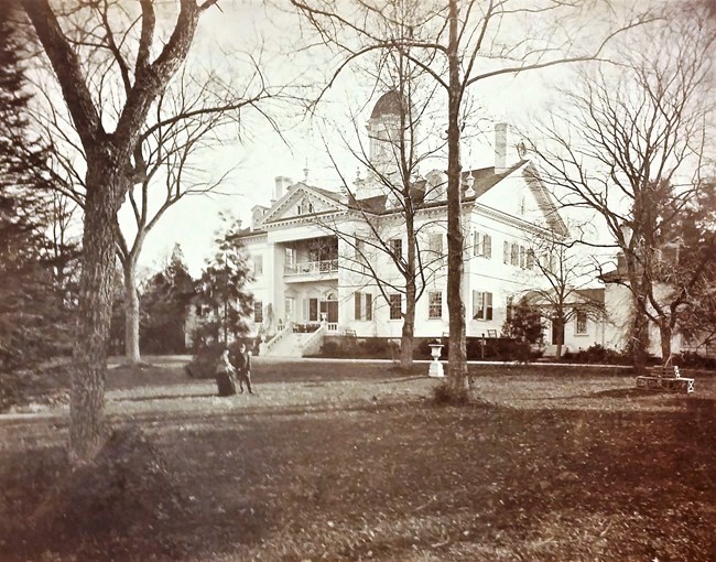 Historic black & white Mansion Photo looking at the north facing side of the mansion, some would say the front of the mansion, but from a slight off center angle. a couple trees in between the camera and the mansion.