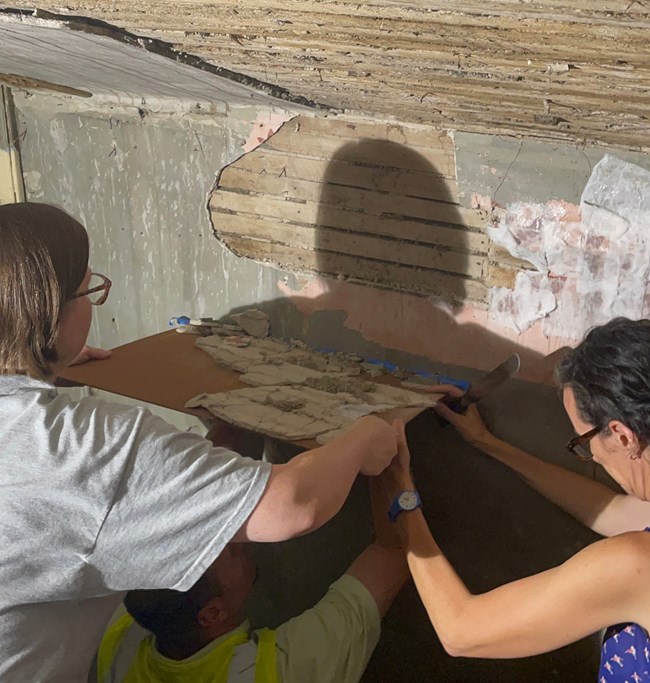 Conservators remove a large section of plaster from a wall using a rigid board for support