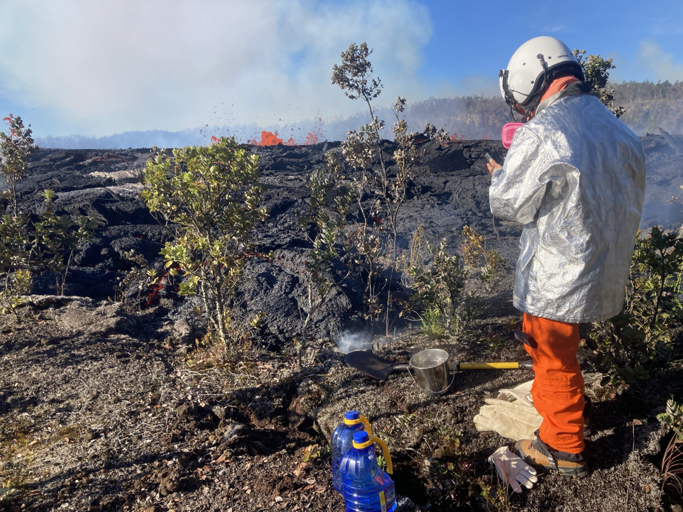 Figure in silver and orange protective gear gathering a lava sample next to dark new lava rock and bright red fountaining lava in the distance.