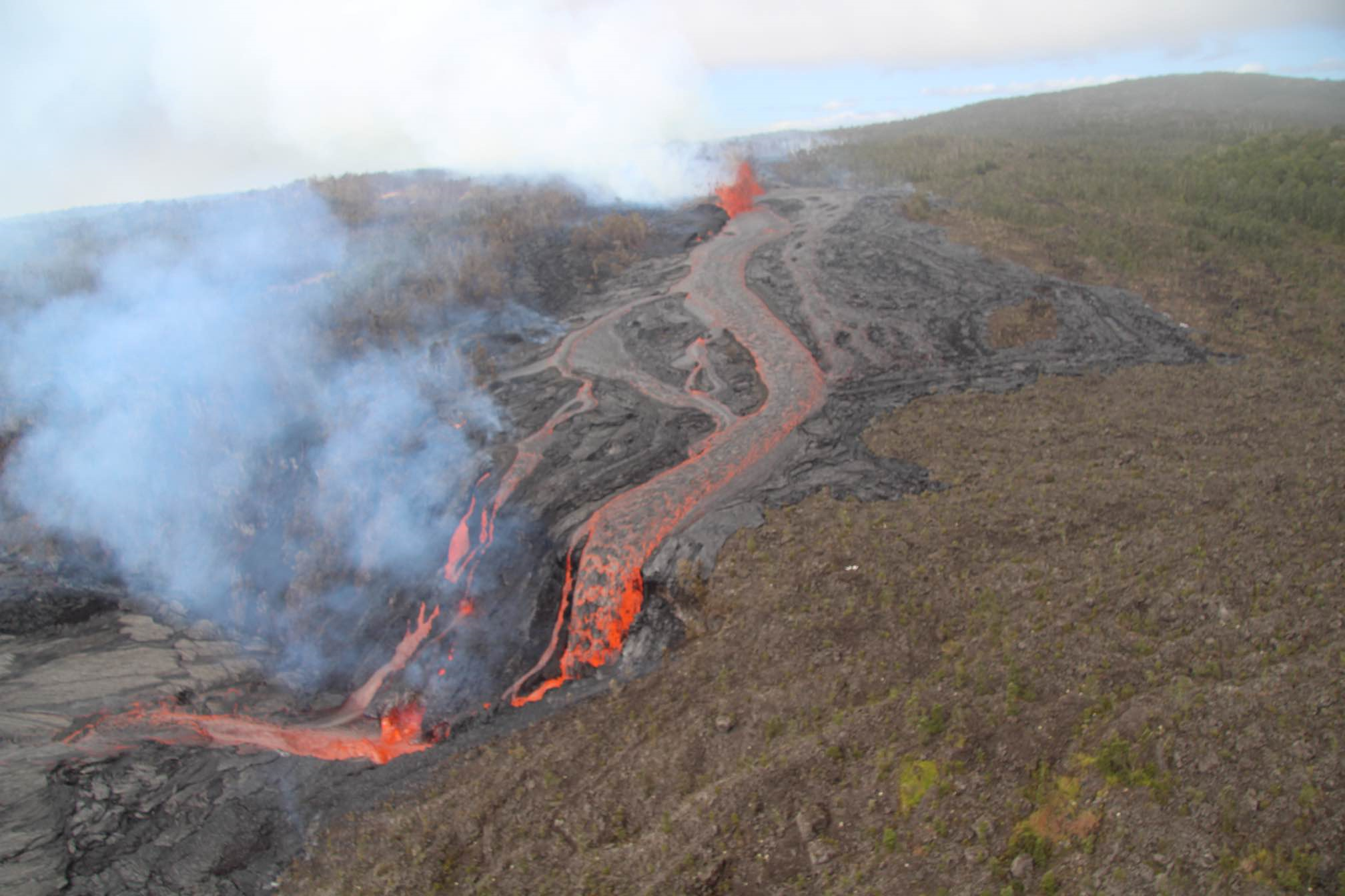 Bright red lava flowing from a fissure high up down a hill and cliff into a crater