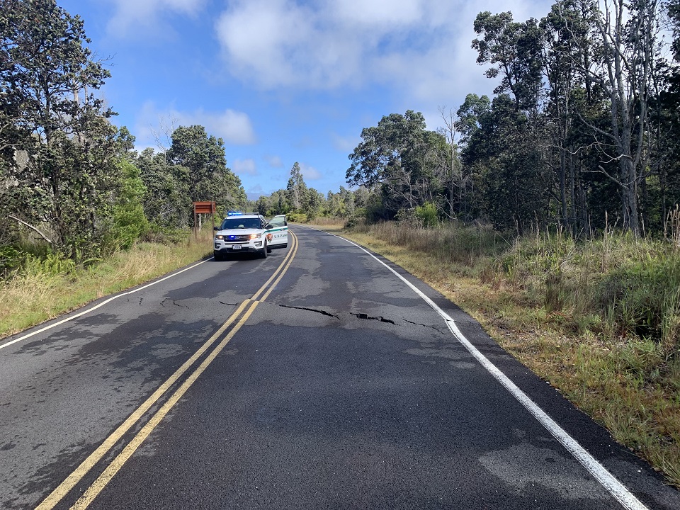 A park ranger vehicle is parked on the road with its door open on the other side of a large crack bissecting a two lane road